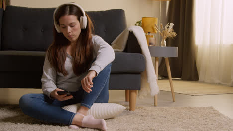 Mujer-Con-Auriculares-Inalámbricos-Sentada-En-El-Suelo-En-Casa-Transmitiendo-Música-Desde-Un-Teléfono-Móvil